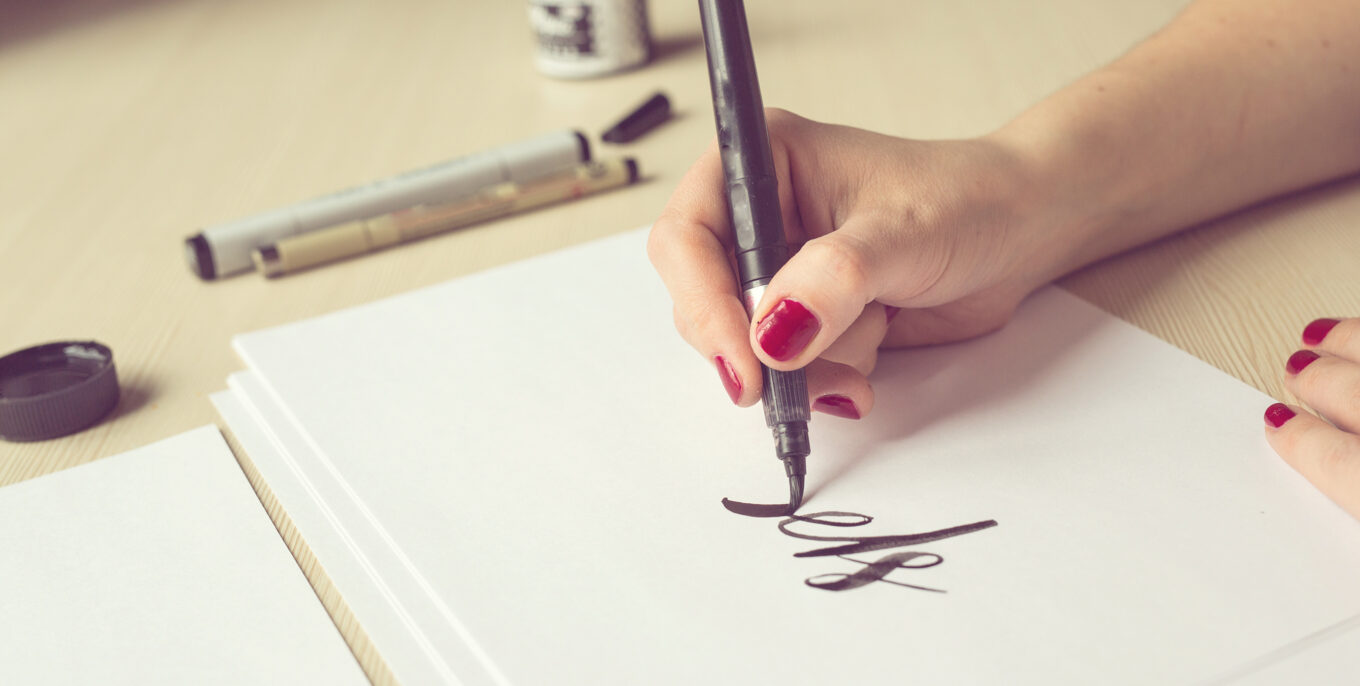 Eine Frau beim Handlettering. Sie hält einen schwarzen Stift und schreibt damit auf weißem Papier. Die Tinte stegt neben ihr auf dem hölzernen Tisch.