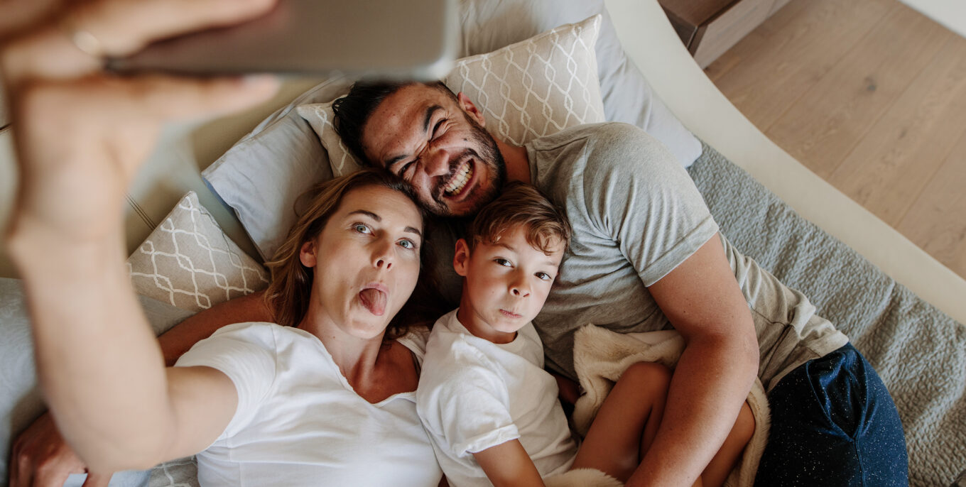 Mutter, Vater, Kind liegen im Bett und machen eine Selfie.