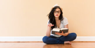 Frau liest Buch. Sie trägt eine Sommerhut, schwarzes, langes haar, eine schwarze Brille, rote Lippen, ein weißes T-Shirt und eine dunkelblaue Jeans. Sie schaut nach unten in das Buch.