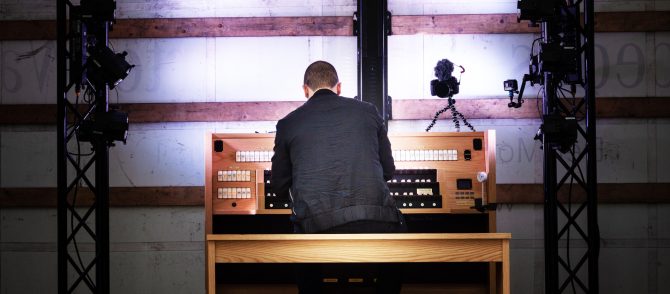 Mann spielt auf einer Orgel. Er ist von hinten zu sehen. Er ist komplett in schwarz gekleidet. Auf der Orgel steht eine Kamera, sowie daneben eine Actionkamera. Der Mann und die Orgel sind gut ausgeleuchtet.