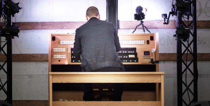 Cameron Carpenter spielt auf einer Orgel. Er ist von hinten zu sehen. Er ist komplett in schwarz gekleidet. Auf der Orgel steht eine Kamera, sowie daneben eine Actionkamera. Der Mann und die Orgel sind gut ausgeleuchtet.