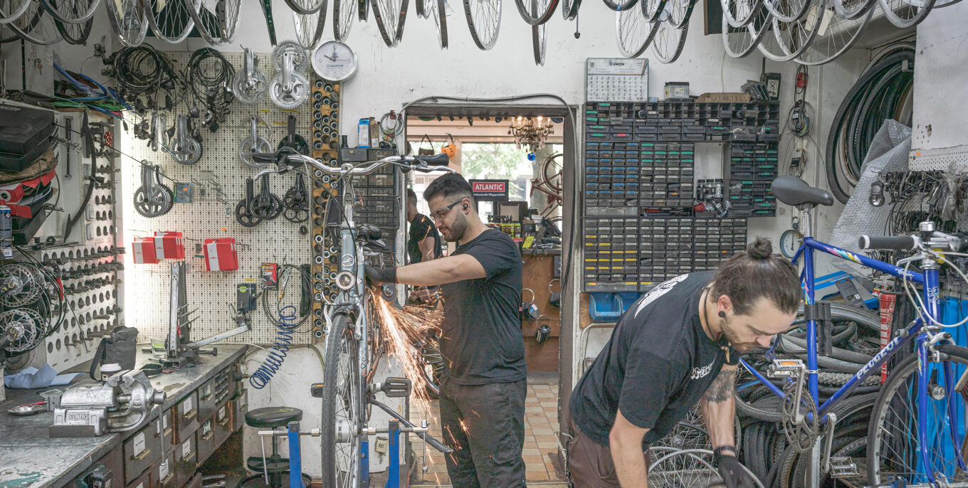 Zwei Männer arbeiten gerade an Fahrrädern in einem Fahrradladen. Um sie herum befinden sich diverse Werkzeuge und Utensilien für Fahrräder.