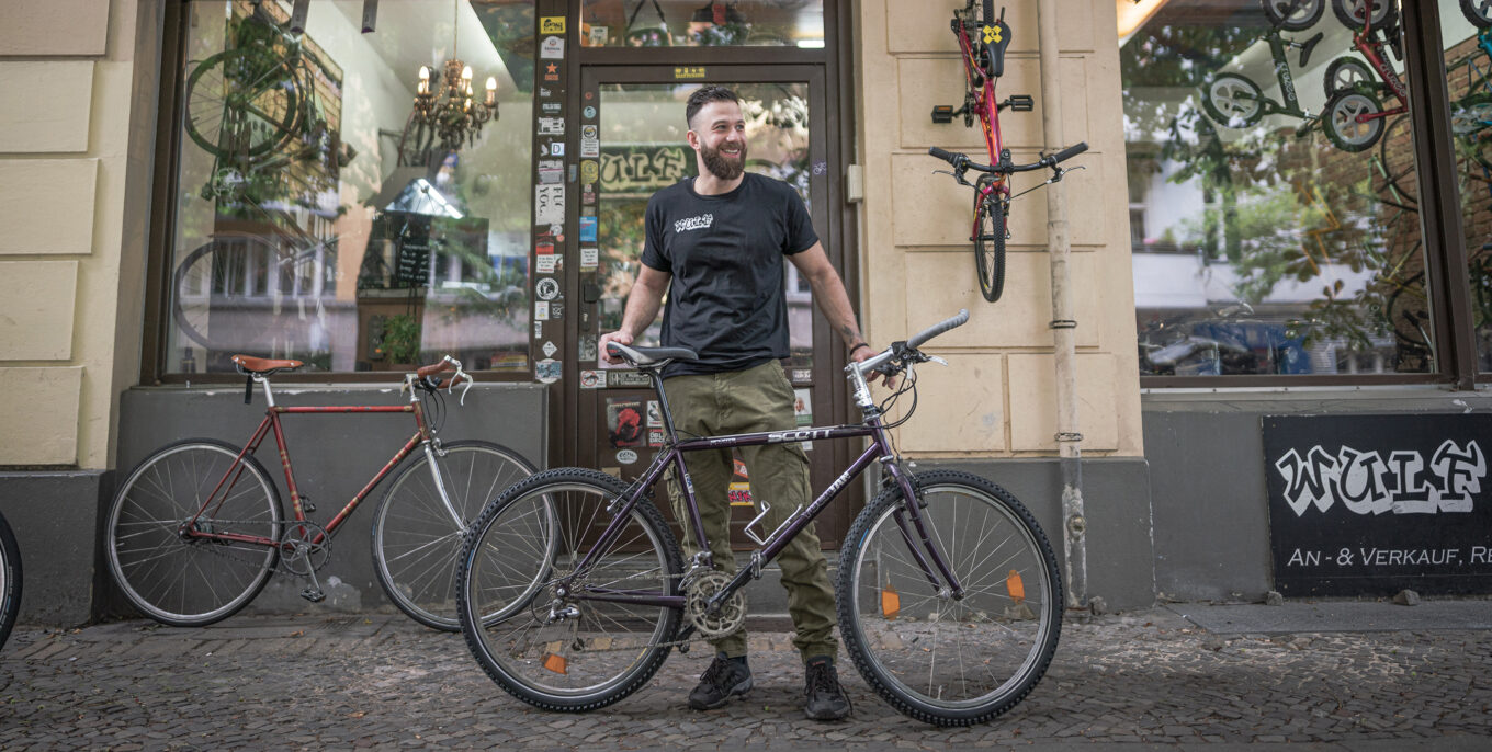 Hicham Safa mit einem Fahrrad schaut seitlich. Er lächelt. Er steht vor einem Fahrradladen.