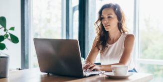 Eine Frau sitzt am Tisch und schaut in ihren Laptop. Neben ihr steht eine Tasse Kaffee, das Sonnenlicht kommt von hinten. Sie trägt ein weißes Top.