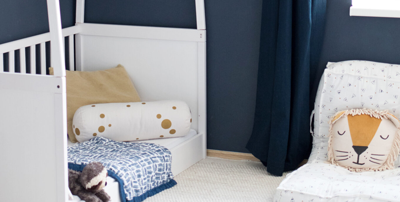 Ein Kinderzimmer mit einem weißen Kinderbettchen. Darauf liegen diverse Kissen, eine Decke und ein Kuscheltier. Daneben sieht man einen Sitz mit einem Kissen in Form eines Löwenkopfes. Die Wand ist dunkelblau.