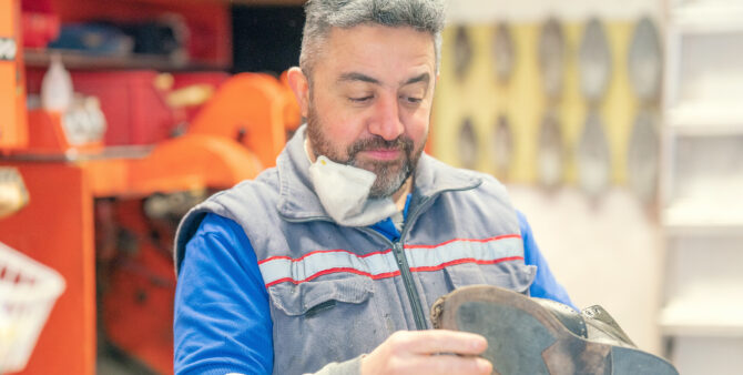 Ibrahim Contur ist konzentriert bei der Arbeit. Er arbeitet gerade an einen Schuh. Er trägt einen blauen Pullover und eine graue Weste. Sein Haar ist angegraut.