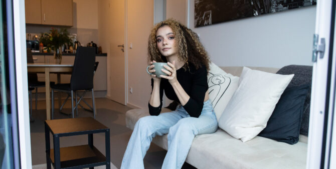 Die Studentin Jasmin trinkt einen Kaffee in ihrer Studentenwohnung. Sie sitzt auf einer Couch, trägt eine hellblaue Jeans und ein schwarzes Top.