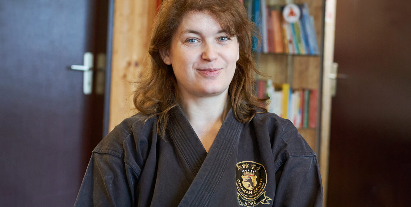 Silvia von Röhl, Karatelehrerin in Spandau, trägt einen dunklen Karateanzug. Man sieht sie im Halbporträt. Im Hintergrund sieht man Bücher und Pokale.