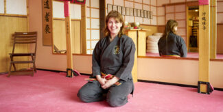 Silvia von Röhl trägt einen dunklen Karateanzug, sie hockt auf dem Fußboden im Übungsraum ihrer Karateschule und lächelt freundlich.