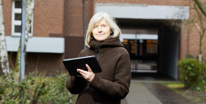 Frau Nowak Janshen steht mit einem iPad in der Hand vor einem Wohnhaus