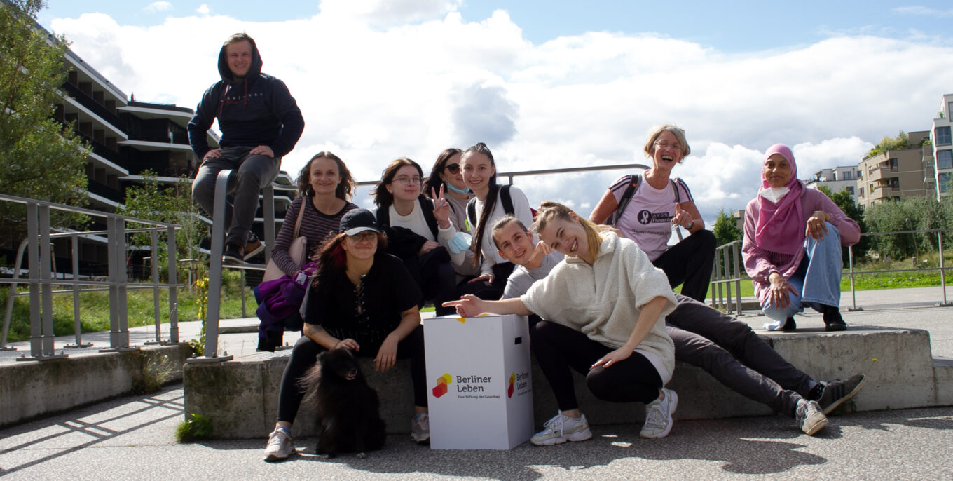 Eine Gruppe Jugendlicher schauen direkt in die Kamera. Sie wirken alle sehr glücklich und versammeln sich um einen Pappkarton mit der Aufschrift "Berliner Leben".