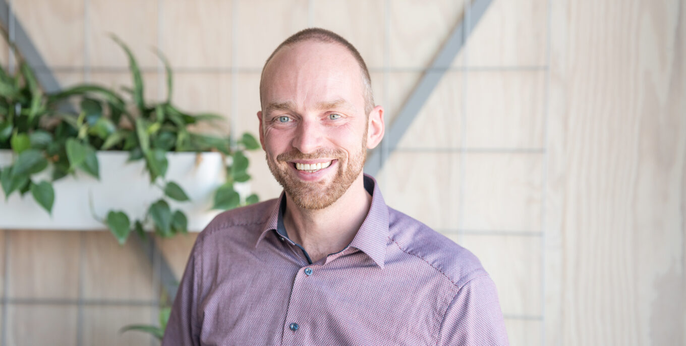 Sören Petzold, Gruppenleiter Technisches Qualitätsmanagement, steht lächelnd vor einer Holzwand mit bepflanzten Blumenkästen. Er trägt ein lilafarbenes Hemd.