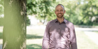 Sören Petzold, Gruppenleiter Technisches Qualitätsmanagement, steht unter einem Baum im Park. Er lächelt in die Kamera, er trägt ein lilafarbenes Hemd. Im Hintergrund sieht man eine große Rasenfläche mit kleinen Wegen.