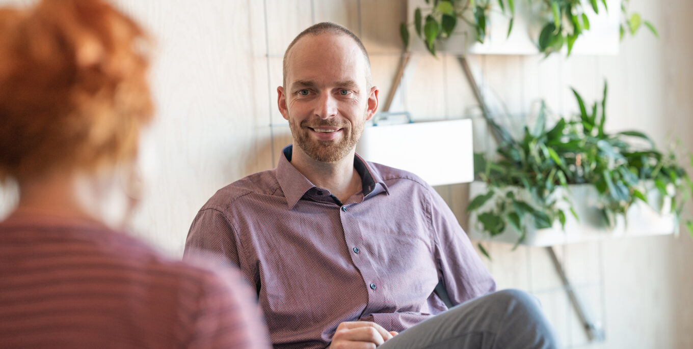 Sören Petzold, Gruppenleiter Technisches Qualitätsmanagement, sitzt vor einer Wand mit bepflanzten Blumenkästen. Ihm gegenüber (Bildvordergrund, unscharf) sitzt eine Frau (von hinten) mit kurzen roten Haaren. Sören Petzold lächelt die Frau an.