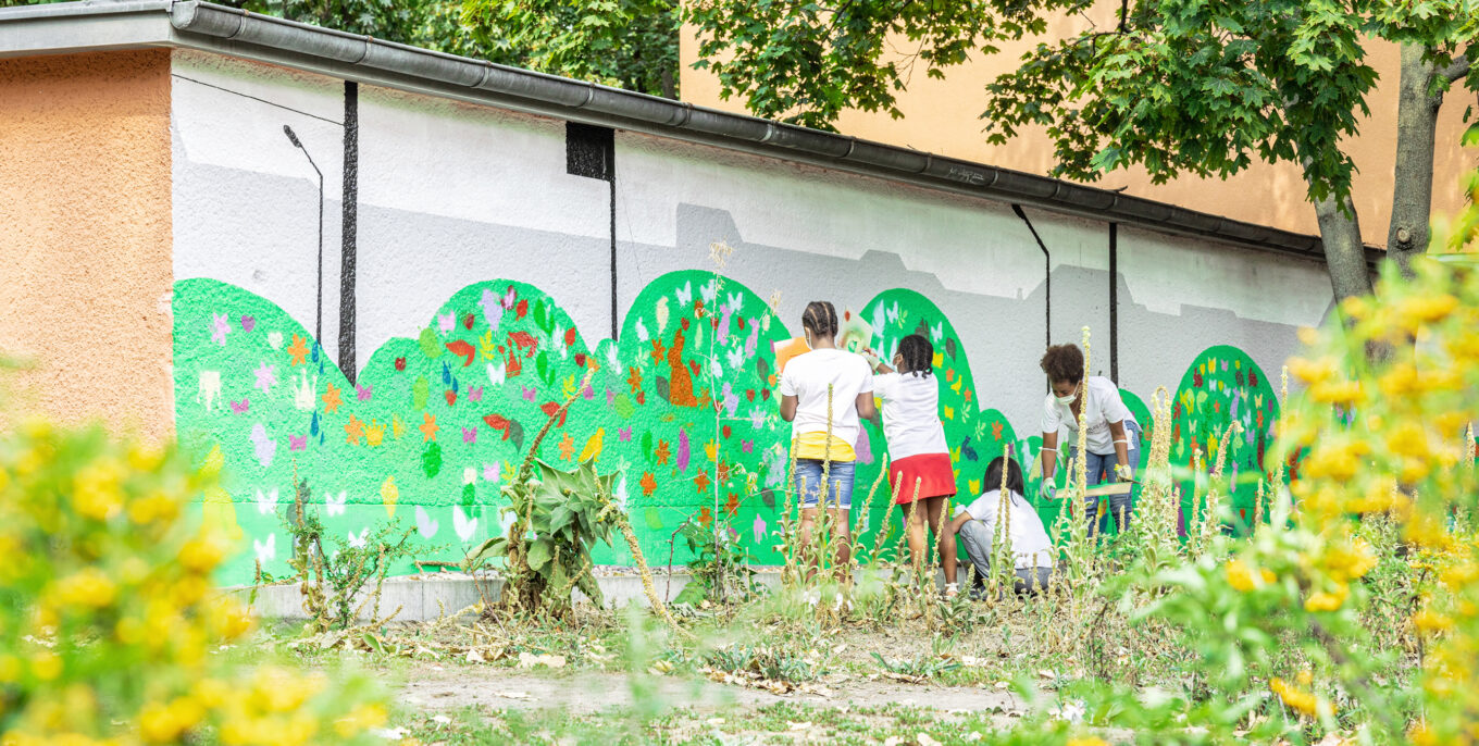 Häuserwand bunt bemalt mit grünen Wiesen und bunten Blumen, Tieren, Herzen und Blättern. Kinder (von hinten) malen an die Häuserwand. Im Vordergrund eine Wiese mit Blumen (unscharf)
