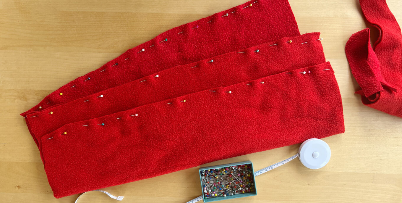 Einzelne rote Stoffteile mit Nadeln fixiert auf Tisch (von oben), daneben steht ein ausgerolltes Maßband und ein Kästchen mit Stecknadeln