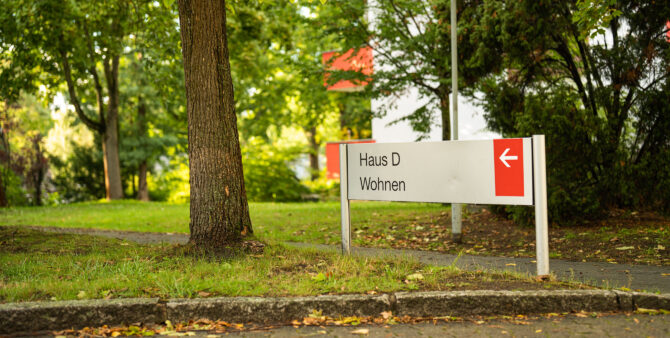 Schild mit Aufschrift: Haus D Wohnen und einem weißen Pfeil auf rotem Untergrund, im Hintergrund und drum herum Wiese und Bäume, teilweise unscharf