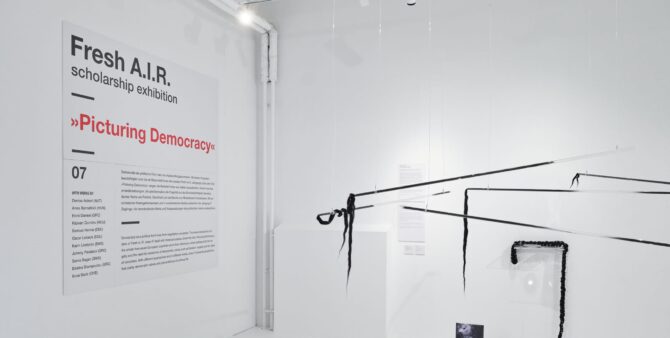 Blick in den Ausstelungsraum, weiße Wände mit einem Plakat und dem Titel der Ausstelung "Picturin Democracy" sowie Ausstellungsgegenstände an den Wänden