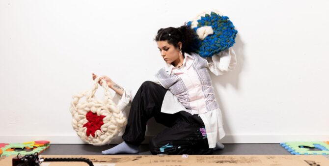 Künstlerlin Sonia Sagan sitzt auf dem Boden und hält zwei gestrickte Gestände in den Händen ihr Blick ist abgewandt Richtugn Boden. Sie trägt eine dunkle Hose, ein weißes Hemd und eine helle Weste. Ihre halblangen schwarzen Harre sind zu einem Zopf gebunden.