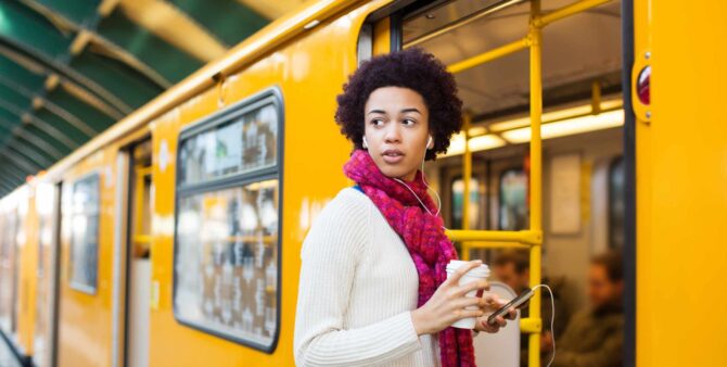 Eine Frau steht vor einer Berliner U-Bahn- Sie trägt Kopfhörer, in der einen Hand hält sie ein Smartphone, in der anderen einen Becher Kaffee.