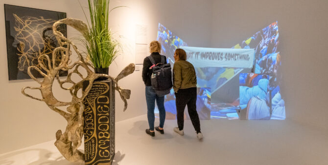 zwei Personen im Ausstellungsraum stehen seitlich vor einer Videoprojektion und schauen auf daneben an der Wand hängenden Erklärtafeln.