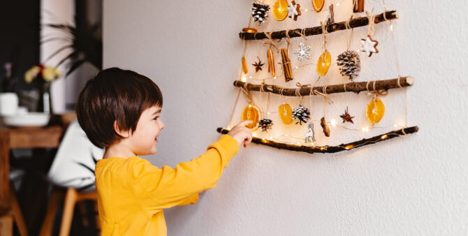 Kleines Kind sieht sich einen handgemachten an der Wand hängenden Weihnachtsbaum aus Ästen und natürlicheren Materialen an.