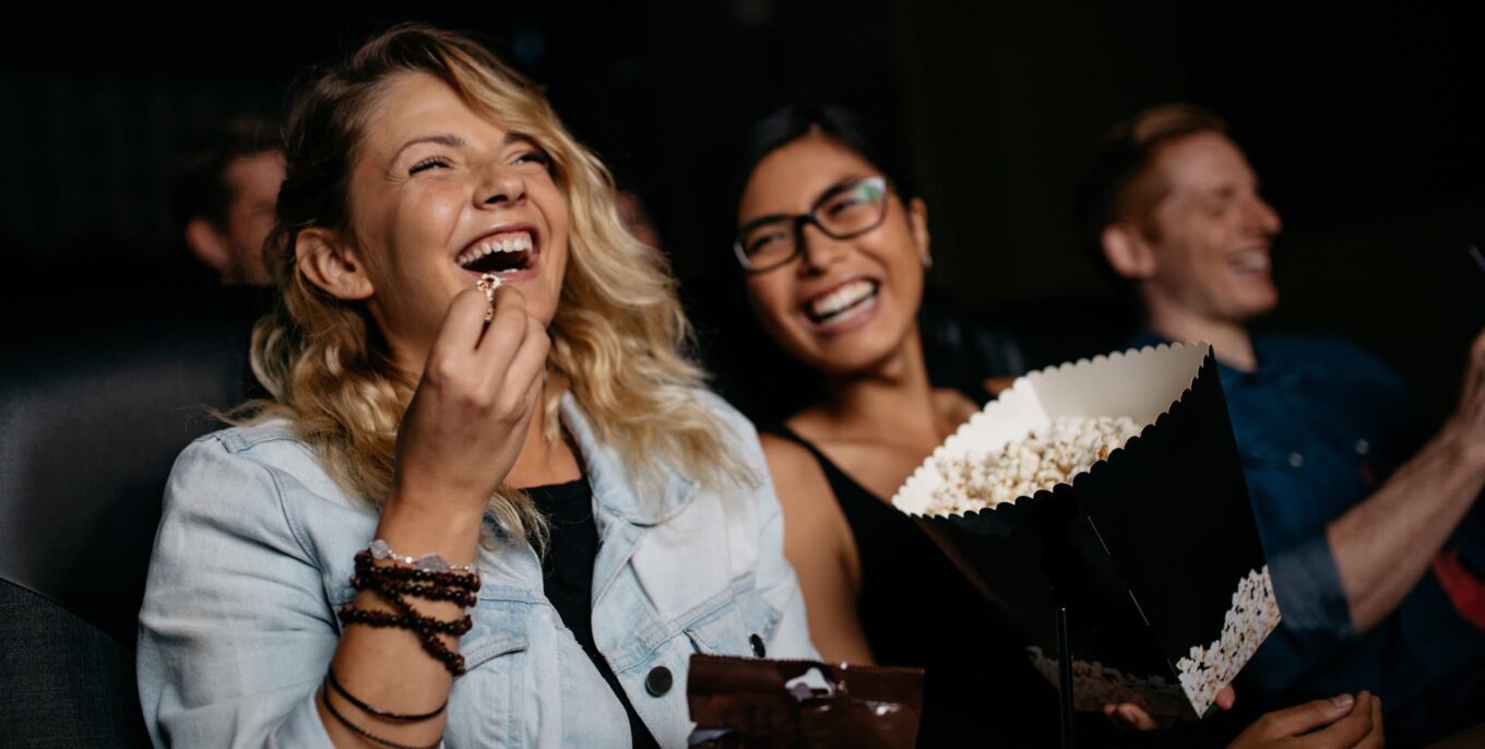 Zwei junge Freuen sitzen nebeneinander im Kino. Sie lachen. Eine isst Popcorn.
