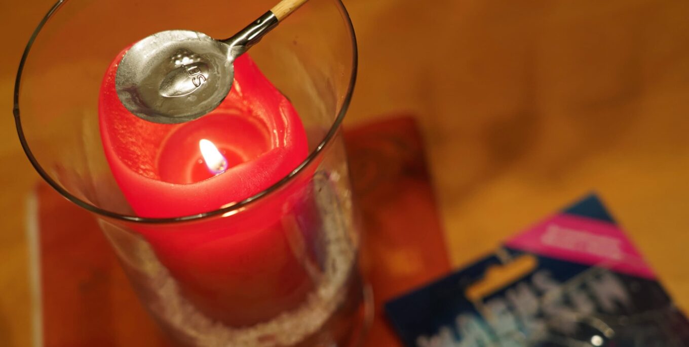 Eine rote Kerze steht in einem Glas. Über die Flamme wird ein kleiner Schmelztiegel gehalten mit einem Wachsstück darin.