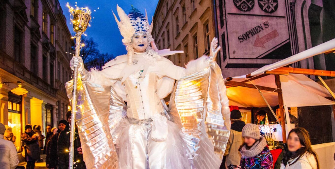 Eine in ein weißes Kostüm gekleide Figur auf Stelzen mit Flügeln läuft über den Umweltweihnachtsmarkt Berlin.