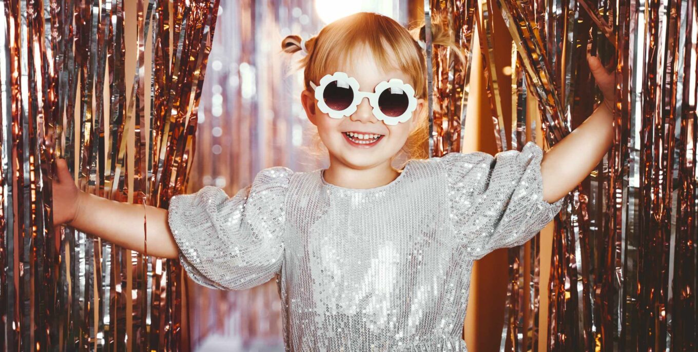 Ein kleines Mädchen mit blonden Zöpfen, Klitzerkleid und einer Sonnenbrille in Blumenform schaut durch einen Vorhang aus Lametta.