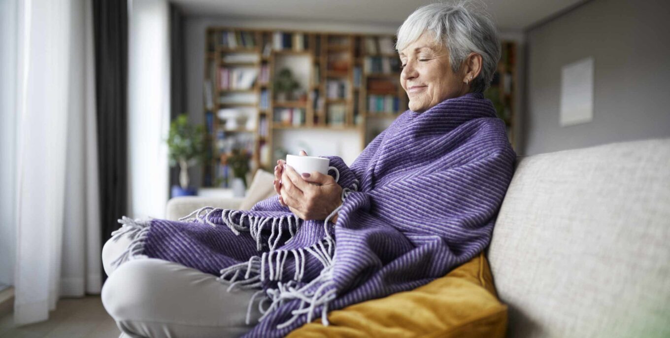 Eine ältere Frau sitzt in eine Decke gehüllt auf einem Sofa. Sie hat die Augen geschlossen und hält eine Tasse in den Händen.
