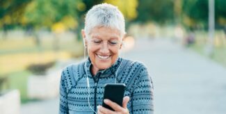Eine ältere Frau in Sportkleidung ist im Freien unterwegs. Sie schaut lächelnd auf ihre Smartphone.