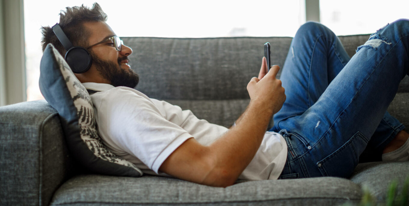 Mann liegt mit Kopfhörern auf einer Couch und schaut auf sein Handy.