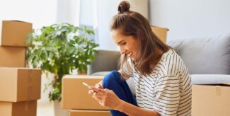 Eine junge Frau sitzt zwischen Umzugskartons und schaut lächelnd auf ihr Handy.