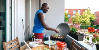 Mann bereitet vegetarische Spieße, Lammfleisch und Würstchen auf elektrischem Grill zu, auf dem Tisch gemischter Salat und Gemüse zum Grillen