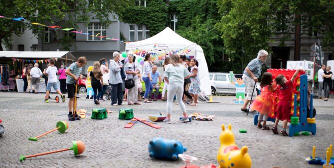 Verschiedene Spielgeräte des Gewobag-Spielmobils liegen ausgebreitet auf einem Platz. Im Hintergrund sind einige Kinder und Erwachsene vor dem Gewobag-Spielmobil zu sehen.