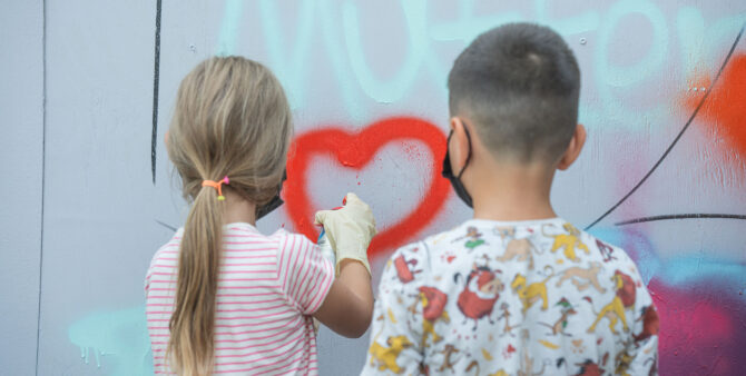 Foto von hinten: Ein Mädchen sprayt während einer Kunstaktion am Jugendclub Heckerdamm ein Herz auf eine Wand, ein Junge schaut zu. Foto: Felix Seyfert