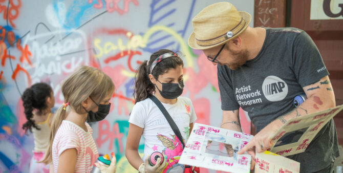 Künstler Christian Rothenhagen spricht mit zwei Mädchen über Motive, die sie im Rahmen einer Kunstaktion am Jugendclub Heckerdamm auf eine Wand sprayen können. Dabei hält er verschiedene Schablonen in der Hand. Foto: Felix Seyfert.