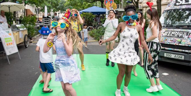 Junge Mädchen mit bunten, übergroßen Brillen, die während des Unartig-Kunstfestivals ausgelassen auf der Straße tanzen. Foto: Nika Kramer