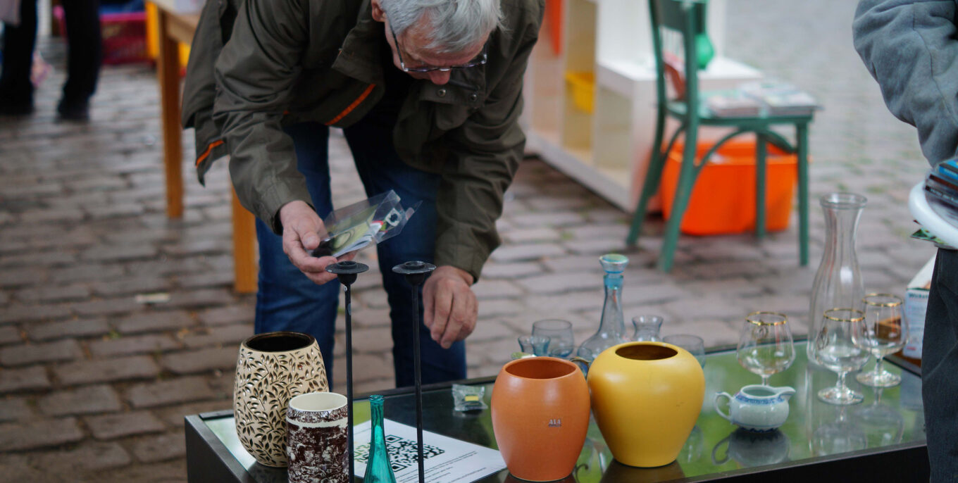 Ein Mann beugt sich auf einem Tausch-Markt über einen Tisch, auf dem Gläser, Vasen und Kerzenständer stehen. Foto: Schön wie wir.