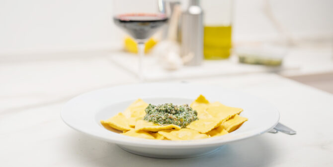Eine Aufnahme von einem Teller gekochten Ravioli mit grünem Pesto. Im Hintergrund steht ein Glas Rotwein. Foto: Felix Seyfert