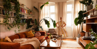 kleine Wohnzimmer: Blick in ein Wohnzimmer, Couch auf der linken Seite, darüber offene Regalbretter mit Büchern, Teppich in der Mitte, durchscheinende Vorhänge, davor steht eine Frau in einem gestreiften Kleid. Foto: Mimi Vollgraf