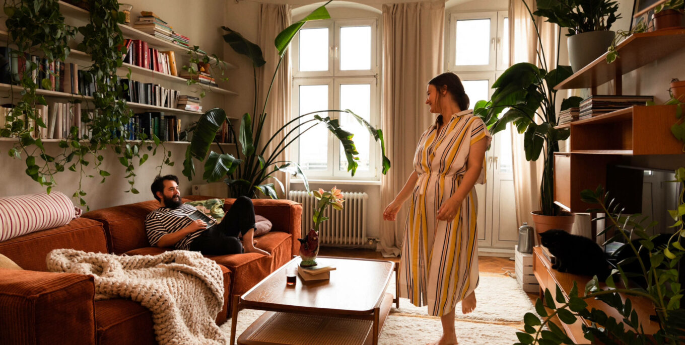 kleines Wohnzimmer: links im Bild steht ein Sofa, auf dem ein Mann sitzt; davor ein Couchtisch; der Mann blickt eine Frau in gestreiftem Klein an, die zwischen Couchtisch und den Regalen auf der rechten Seite entlangläuft. Foto: Mimi Vollgraf