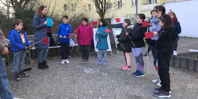 Eine Gruppe Kinder bei der Begehung des ursprünglichen Spielplatzes am Blasewitzer Ring mit Gewobag-Mitarbeiterin Ana Stoeckermann. Die Kinder halten Holzstäbe mit einem daran befestigten roten Fähnchen in den. Foto: Gewobag