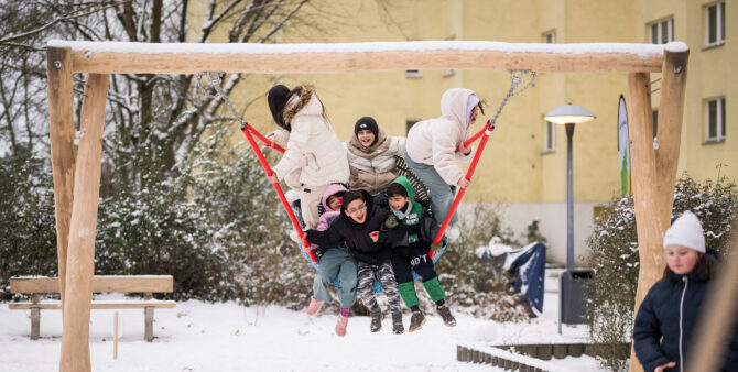 Eine Gruppe Kinder schwingt lachend auf einer Netzschaukel auf einem verschneiten Spielplatz. Foto: Moritz Eden/City-Press GmbH