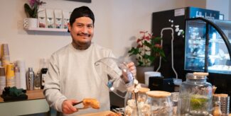 Ein Mann mit umgedrehten Basecap steht hinter dem Tresen eines Cafés und hält in einer Zange ein Gebäckstück. Foto: Ralph Maak