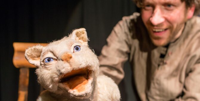 Ein heller Stofftier Kater scheint in die Kamera zu brüllen, dahinter ist ein Puppenspieler im grau-braunen Hemd zu erkennen. Foto: Theater Zitadelle / Klaus Zinnecker