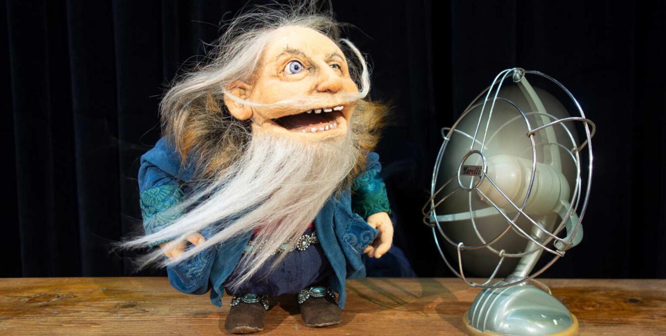 Eine Puppe mit langem grauen Bart, fusseligen grauen Haaren und breitem Mund steht auf einem Tisch neben einem Tischventilator auf der Bühne eines Kindertheaters. Foto: Theater Zitadelle / Vesuv