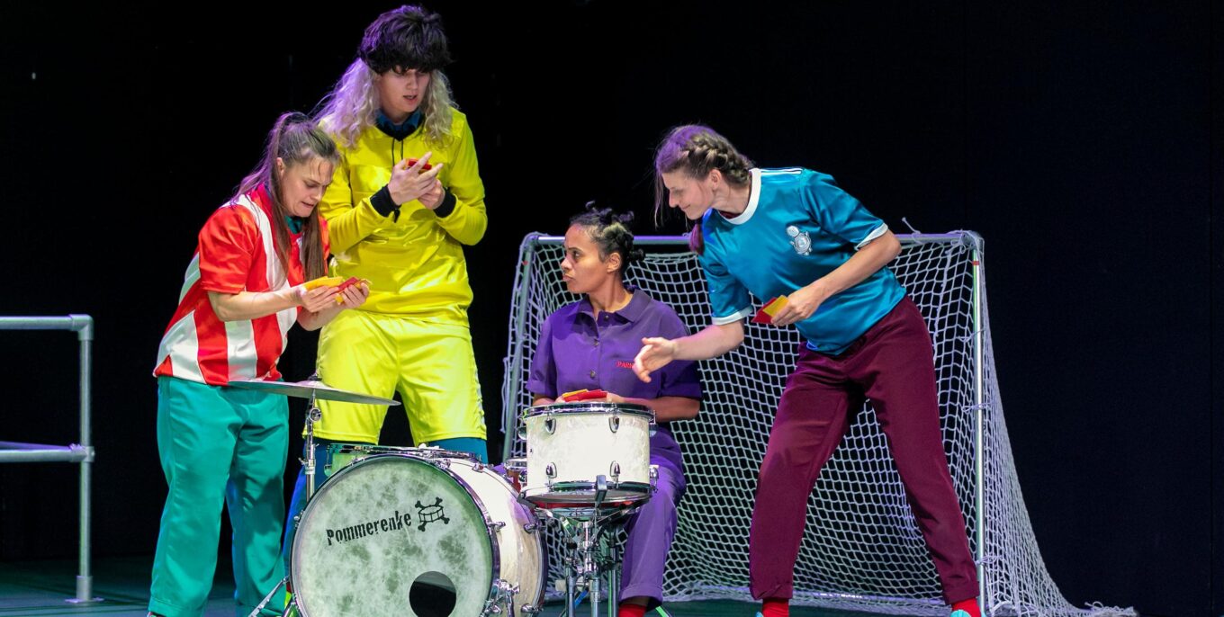 Auf der Bühne eines Kindertheaters in Berlin befinden sich vier Personen in bunten Klamotten vor einem kleinen Fußballtor. Eine Frau in der Mitte sitzt am Schlagzeug, die anderen scheinen über gelbe und rote Karten zu diskutieren. Foto: Theater an der Parkaue / David Baltzer