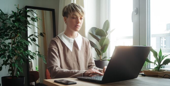 Eine junge Frau mit kurzen blonden Haaren sitzt in einer Wohnung an einem Tisch vor einem Fenster und arbeitet an einem Laptop. Foto: Christian Rückert